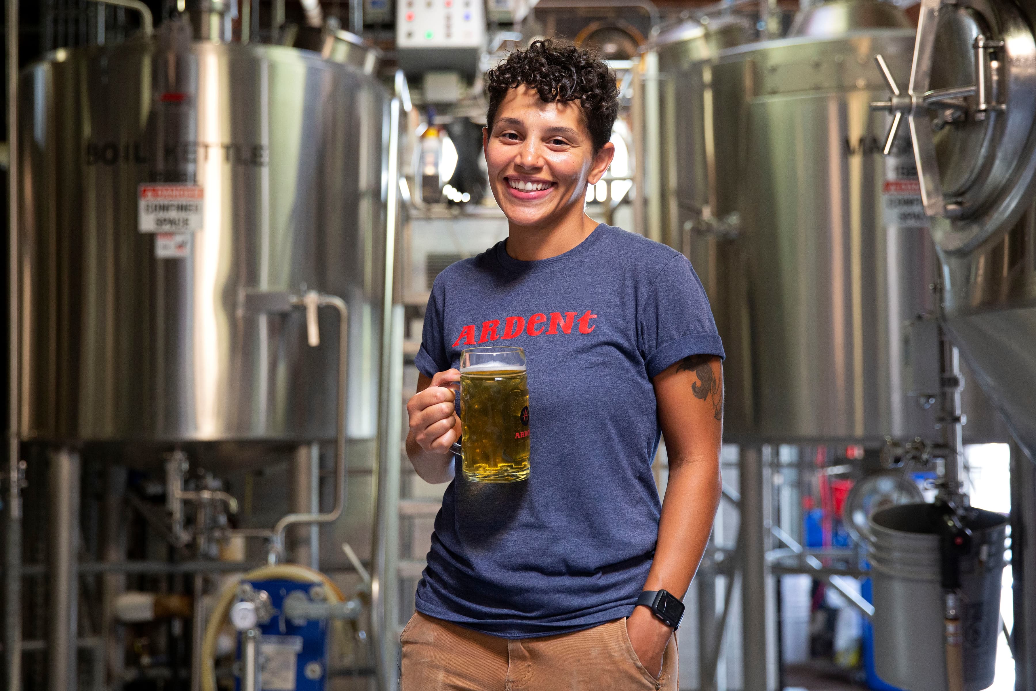 Woman smiling holding stein of beer inside brewery surrounding by brewing tanks