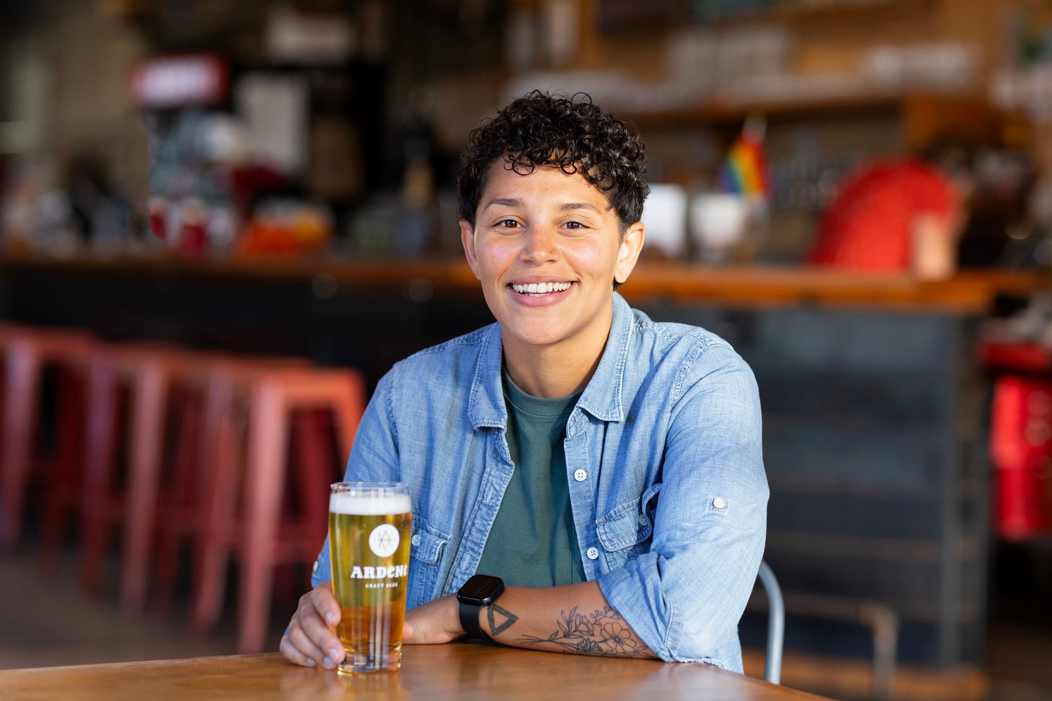 Young woman wearing denim with tattooed arm holding a pint of beer sitting in a taproom