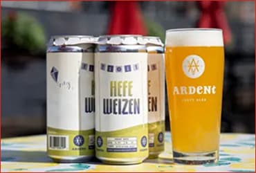 4-pack of white 16oz cans of Hefeweizen next to 16oz glass of cloudy, golden beer