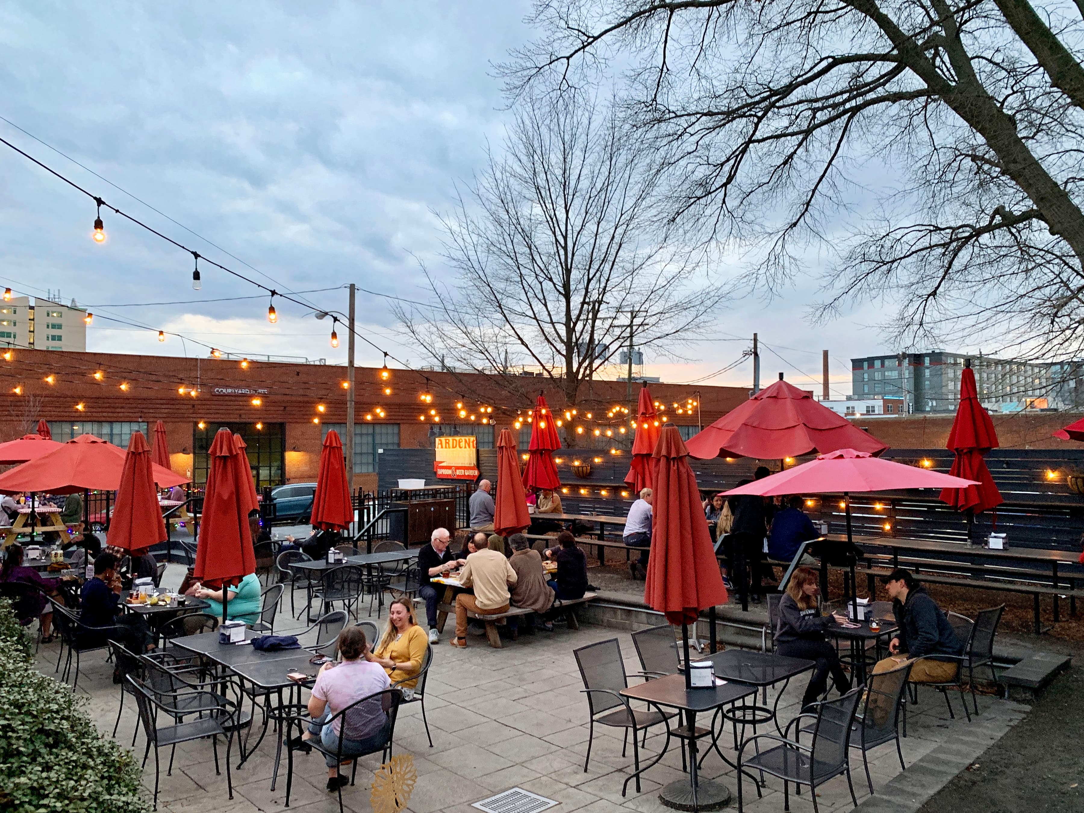String lights over a beer garden patio with red umbrellas and people at tables