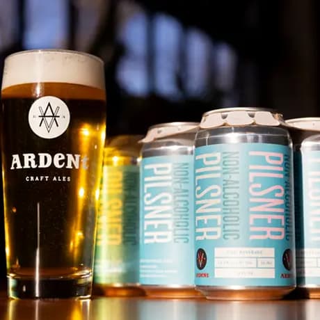 6-pack of pale blue 12oz cans of Non-Alcoholic Pilsner next to tall 16oz glass full of clear golden lager