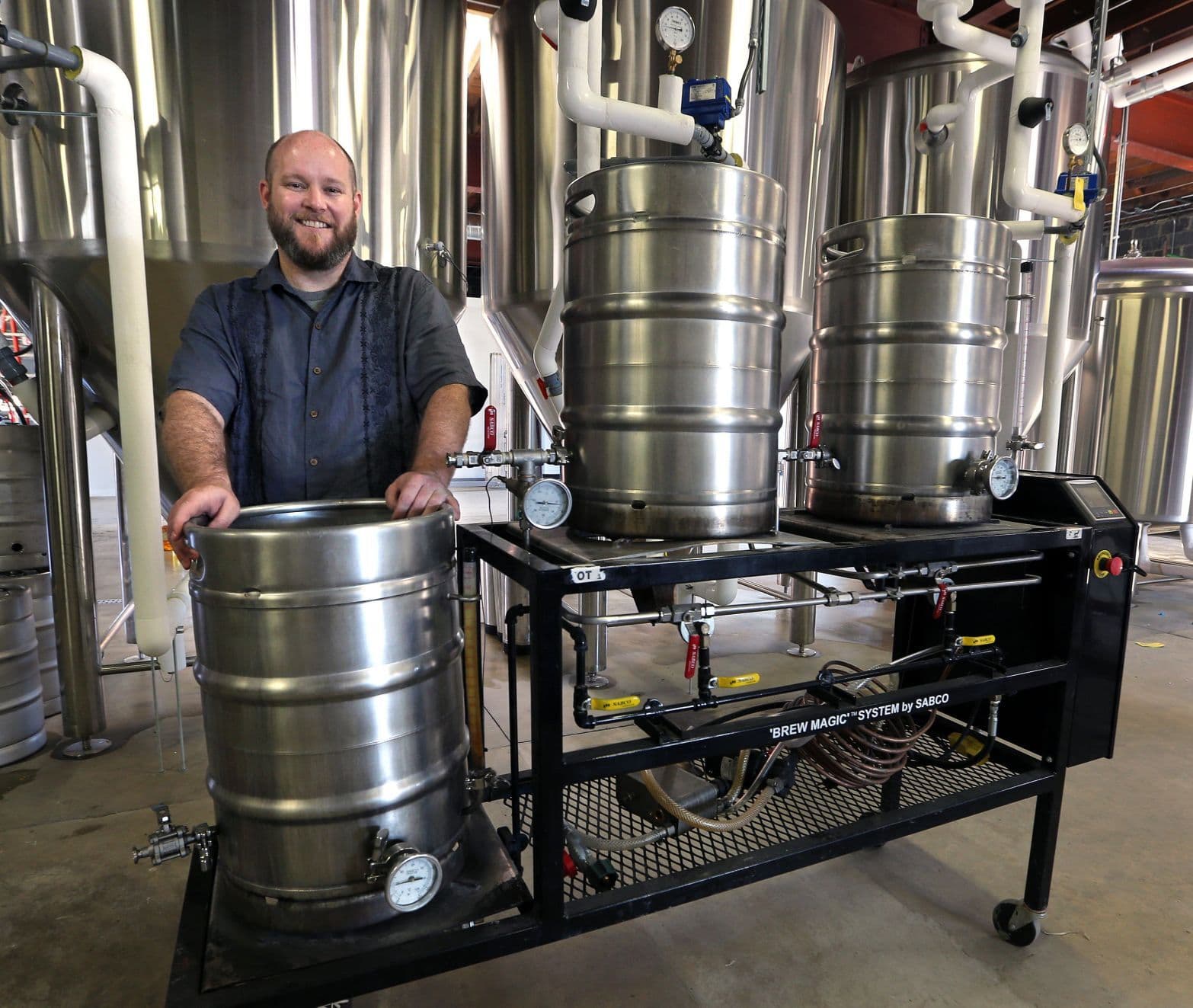 Man wearing work shirt standing with a small brewing system of 3 kegs