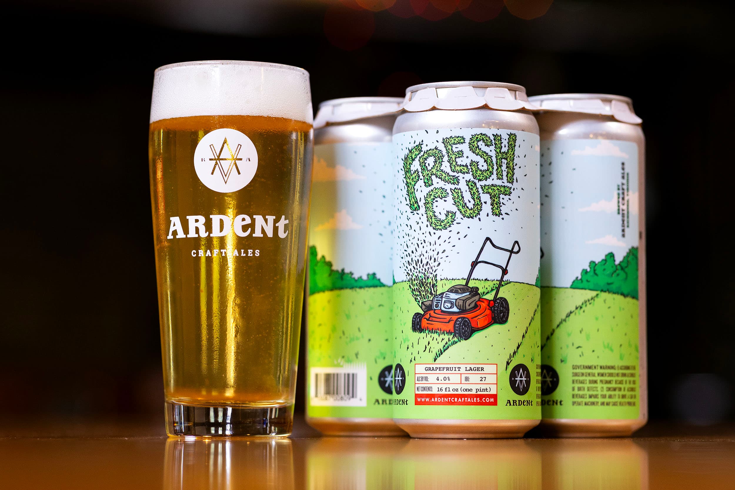 4-pack of pale blue 16oz cans of Fresh Cut grapefruit lager next to tall 16oz glass of clear golden beer