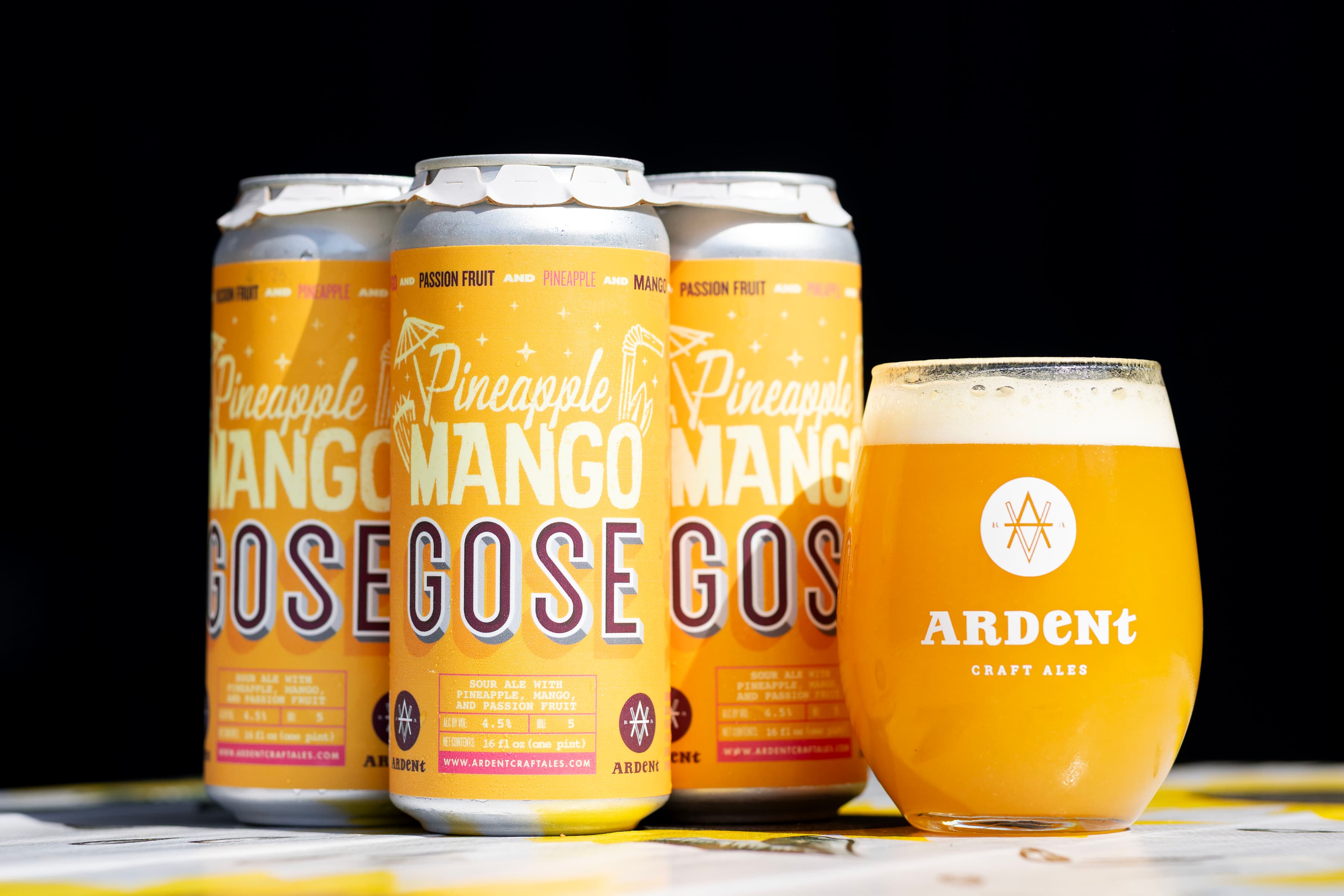 4-pack of orange 16oz cans of Pineapple Mango Gose next to stemless 12oz glass of cloudy, golden beer