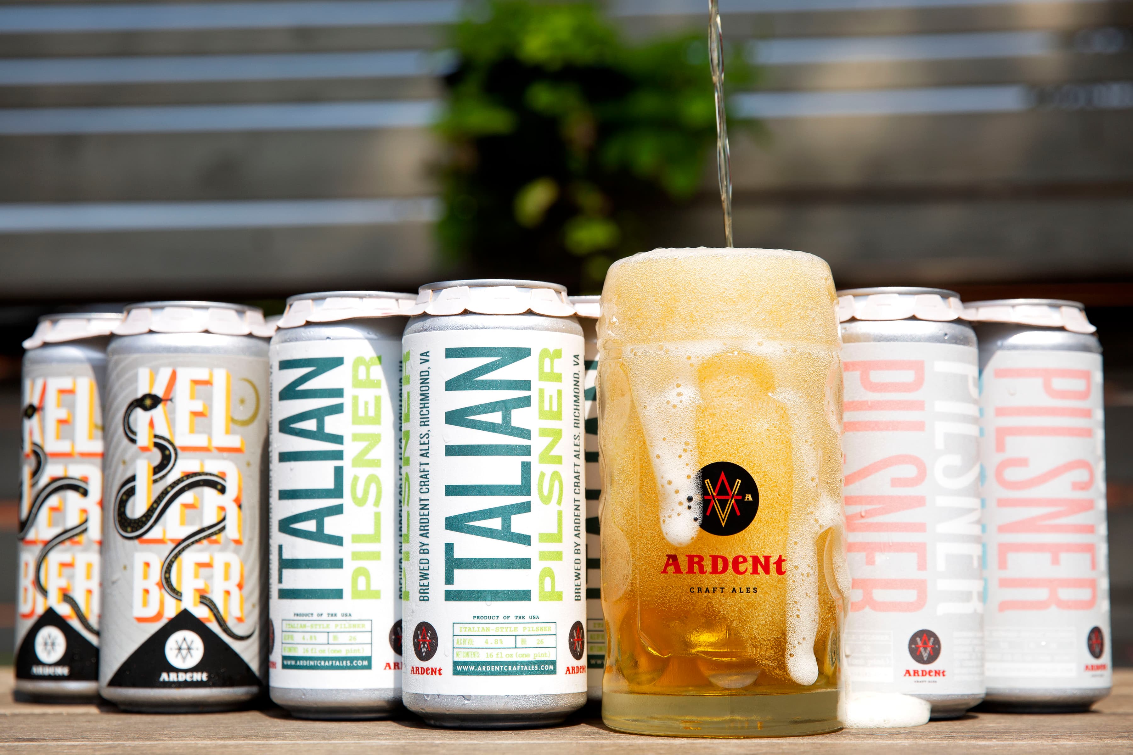 Beer being poured into an overflowing stein in front of various 4-packs of pilsner cans